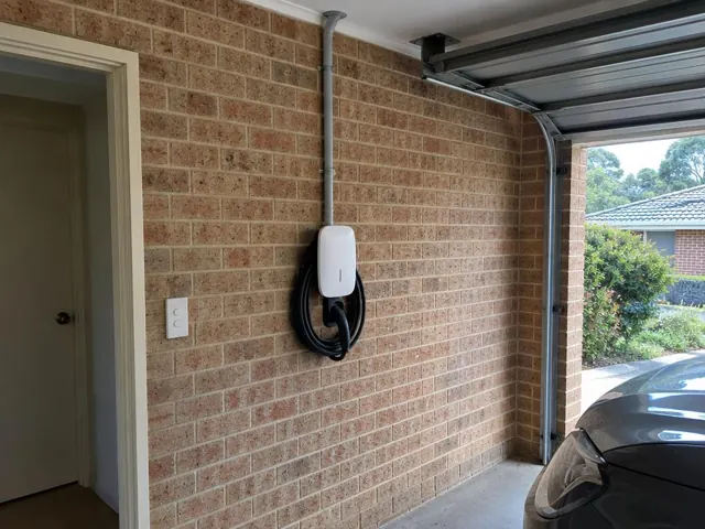 Wall-mounted EV charger on a brick garage wall beside a doorway and open garage door in Kincumber